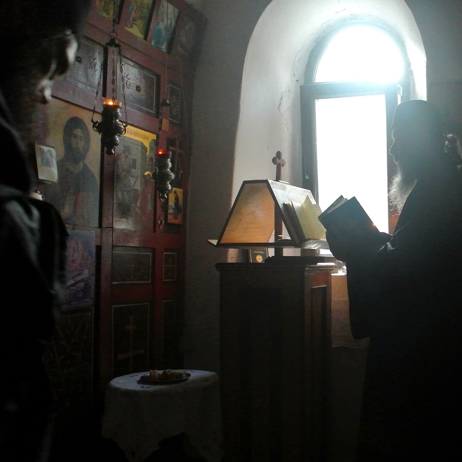 A chapel interior with icons and hanging lamps, and a monk reading beside a sunlit window